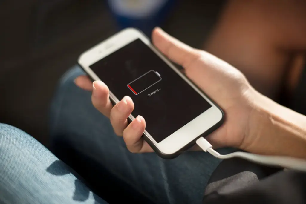 A close-up of a person holding a white smartphone connected to a lightning cable, where the screen shows a critical low battery indicator and reads 'Charging', indicating a worn out cell that needs a professional battery replacement.