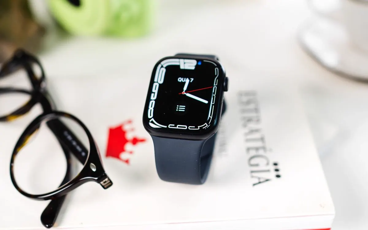 A smartwatch with a dark band resting on a notebook beside a pair of glasses