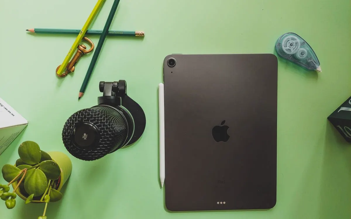 A dark grey iPad lying flat on a green desk beside pencils and a lens