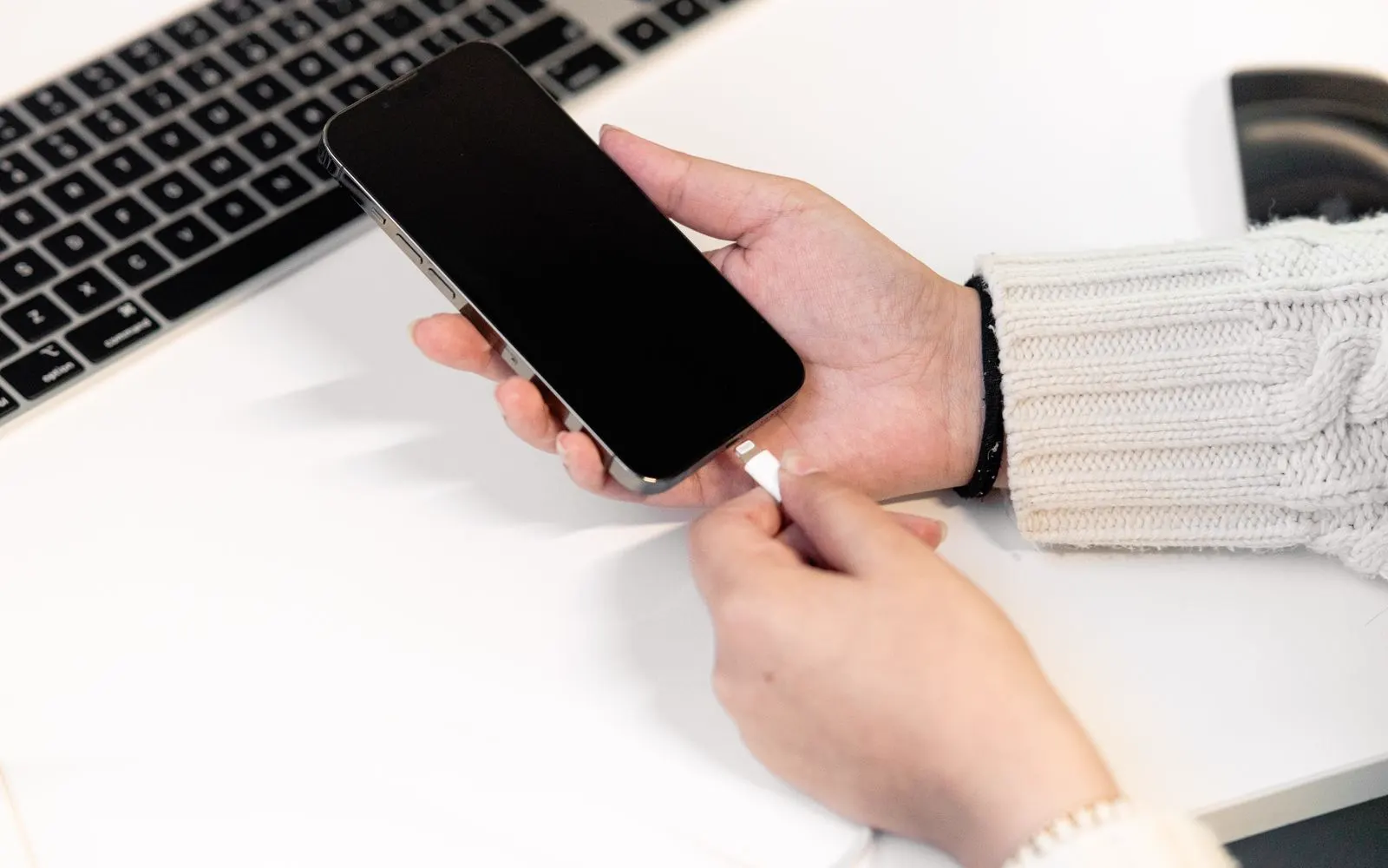 A person plugging a phone into a charger at a desk beside a laptop