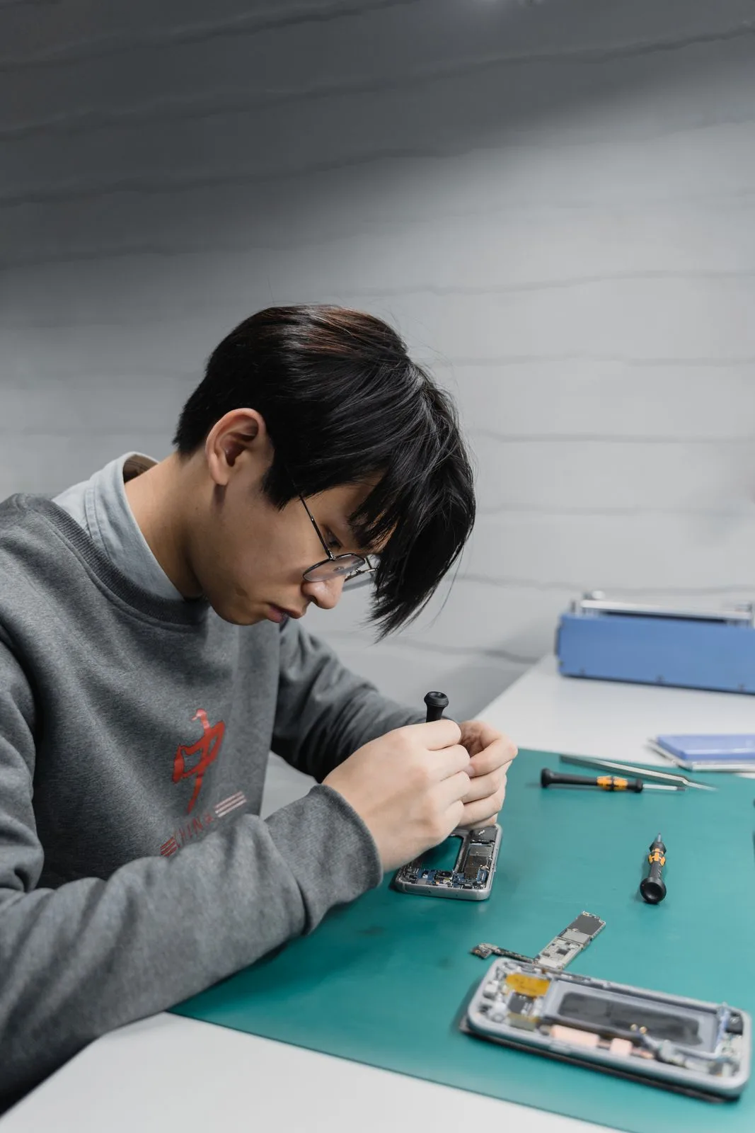 A technician working on the opened internals of a smartphone at a bench, tools and a second device laid out beside it