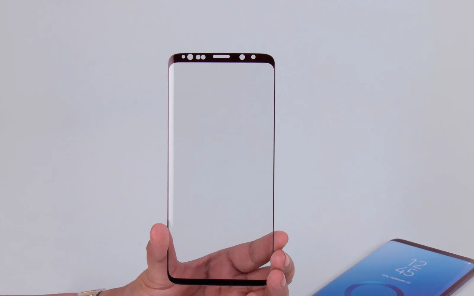 A hand holding a curved tempered-glass screen protector beside a Galaxy phone